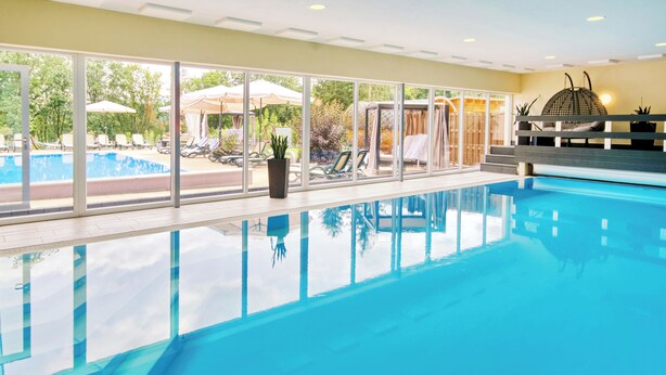 Une piscine intérieure avec des parois en verre transparent, des chaises longues et des parasols à l’extérieur.