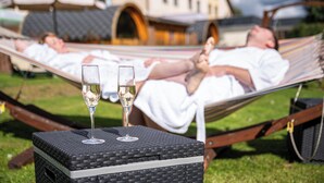 Une personne allongée dans un hamac, sur une pelouse, avec une table devant elle et deux verres de vin.