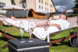 A person lying in a hammock on a grassy area with a table and two glasses of wine in front.