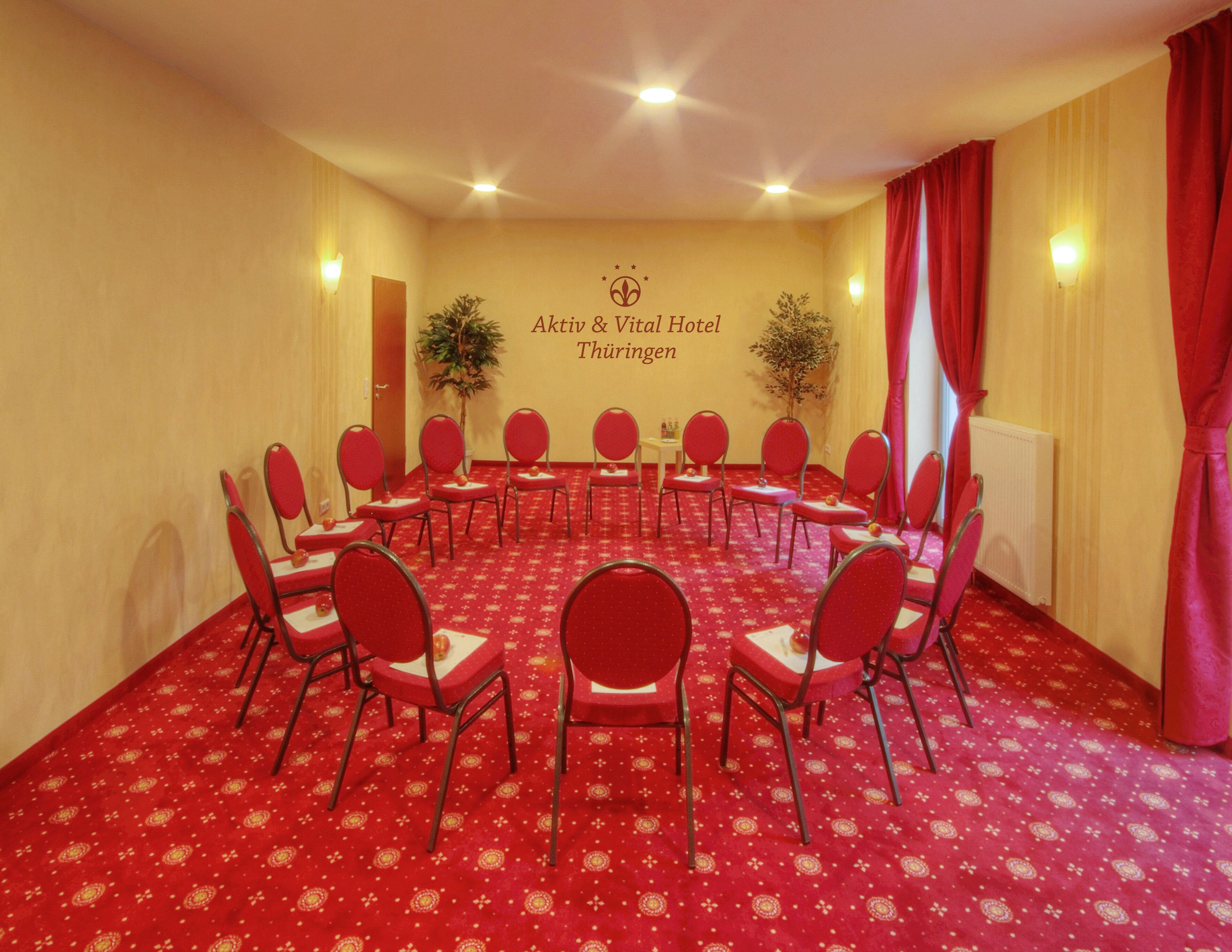 Ein Konferenzraum mit roten, kreisförmig angeordneten Stühlen, einem Teppichboden und einer Wand mit dem Namen des Hotels.