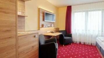 Ein Hotelzimmer mit einem Holzschrank, einem Schreibtisch mit Flachbildfernseher, zwei schwarzen Sesseln, einem roten Teppich mit Muster und einem großen Fenster mit durchsichtigen Vorhängen.