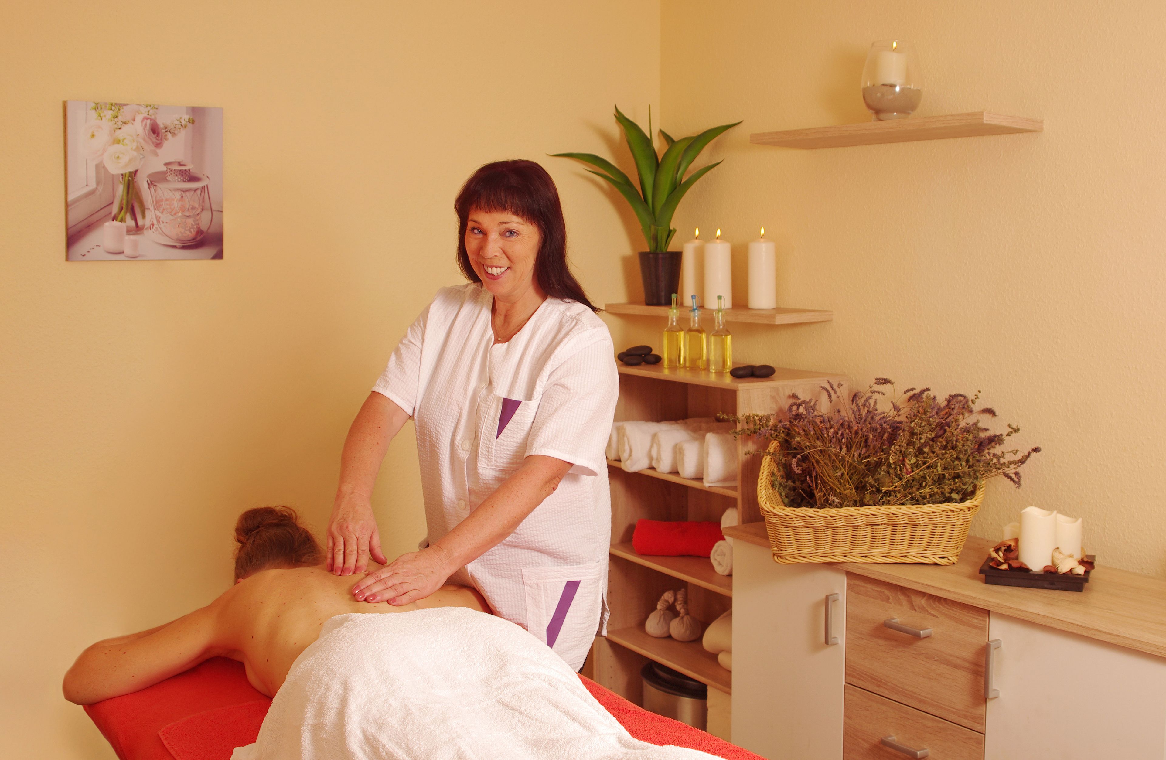 A massage therapy room with a client lying on a red massage table, a therapist standing beside them, and a shelf with candles, a potted plant, and a basket of dried flowers.
