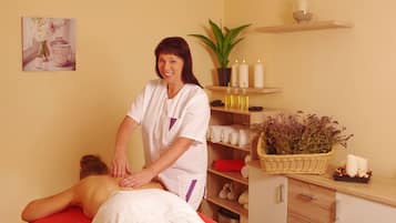 A massage therapy room with a client lying on a red massage table, a therapist standing beside them, and a shelf with candles, a potted plant, and a basket of dried flowers.