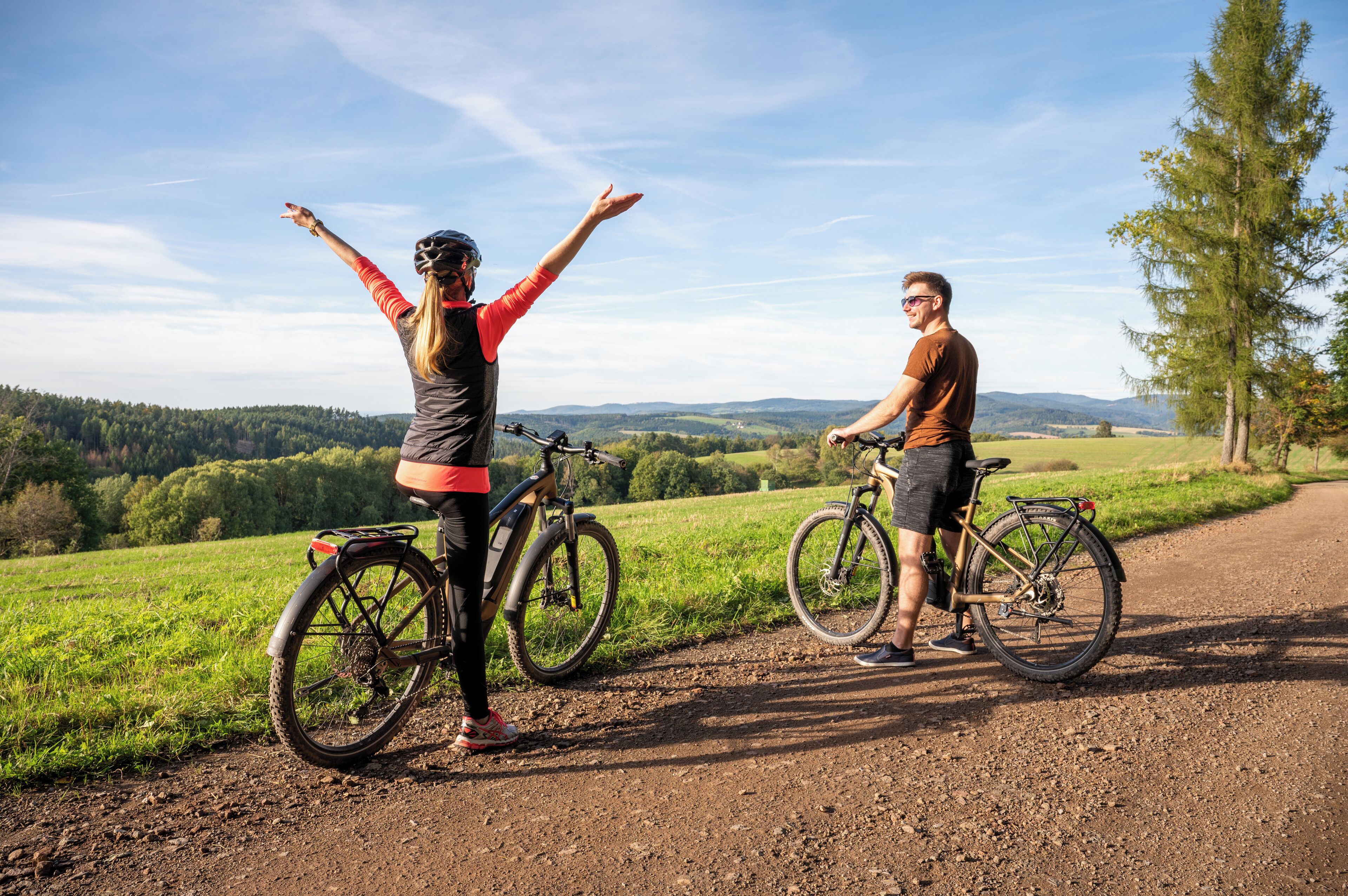 Two people with bicycles on a dirt path with a scenic view of a forest and hills in the background.