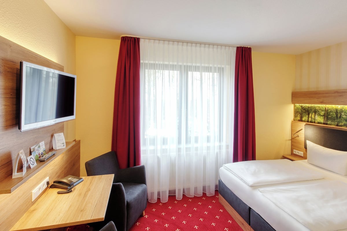 Une chambre d’hôtel comprenant un lit, un bureau avec un téléphone, une chaise, un téléviseur fixé au mur et une fenêtre avec des rideaux légers.
