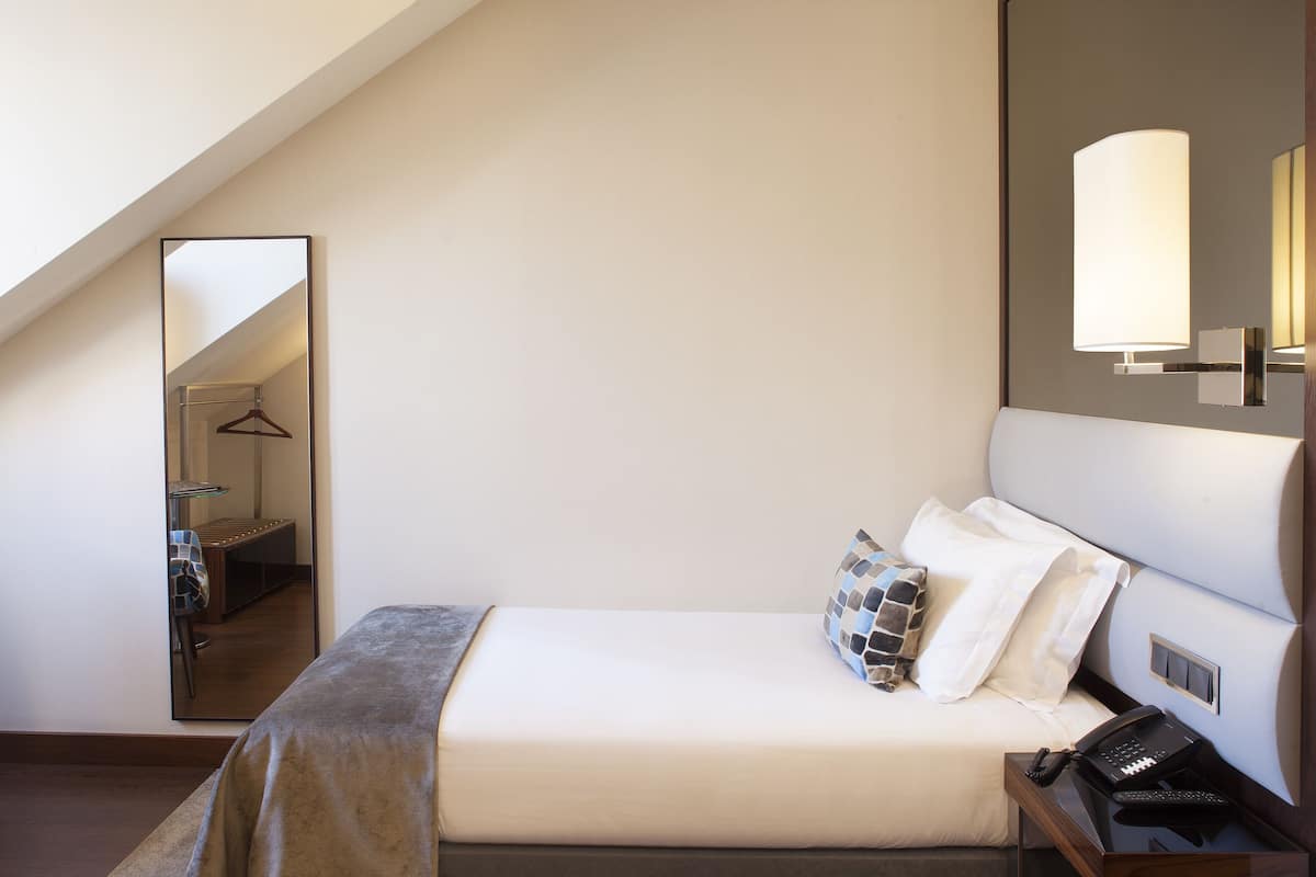 Habitación de hotel con una cama, una mesita de noche con teléfono, una lámpara de pared y un espejo que refleja un pasillo con perchero.