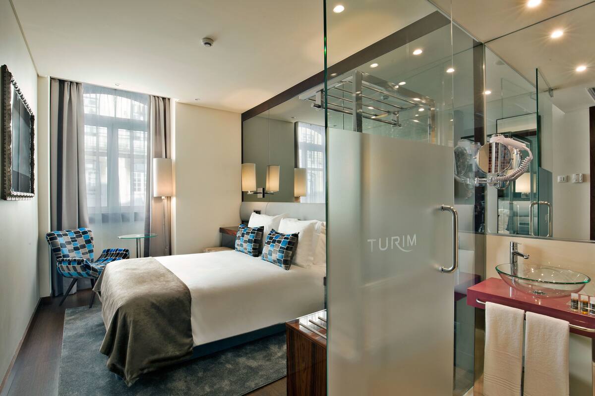 Habitación de hotel moderna con una cama grande, un box de ducha con paredes de vidrio, un lavamanos de cristal y una zona de estar con una silla.