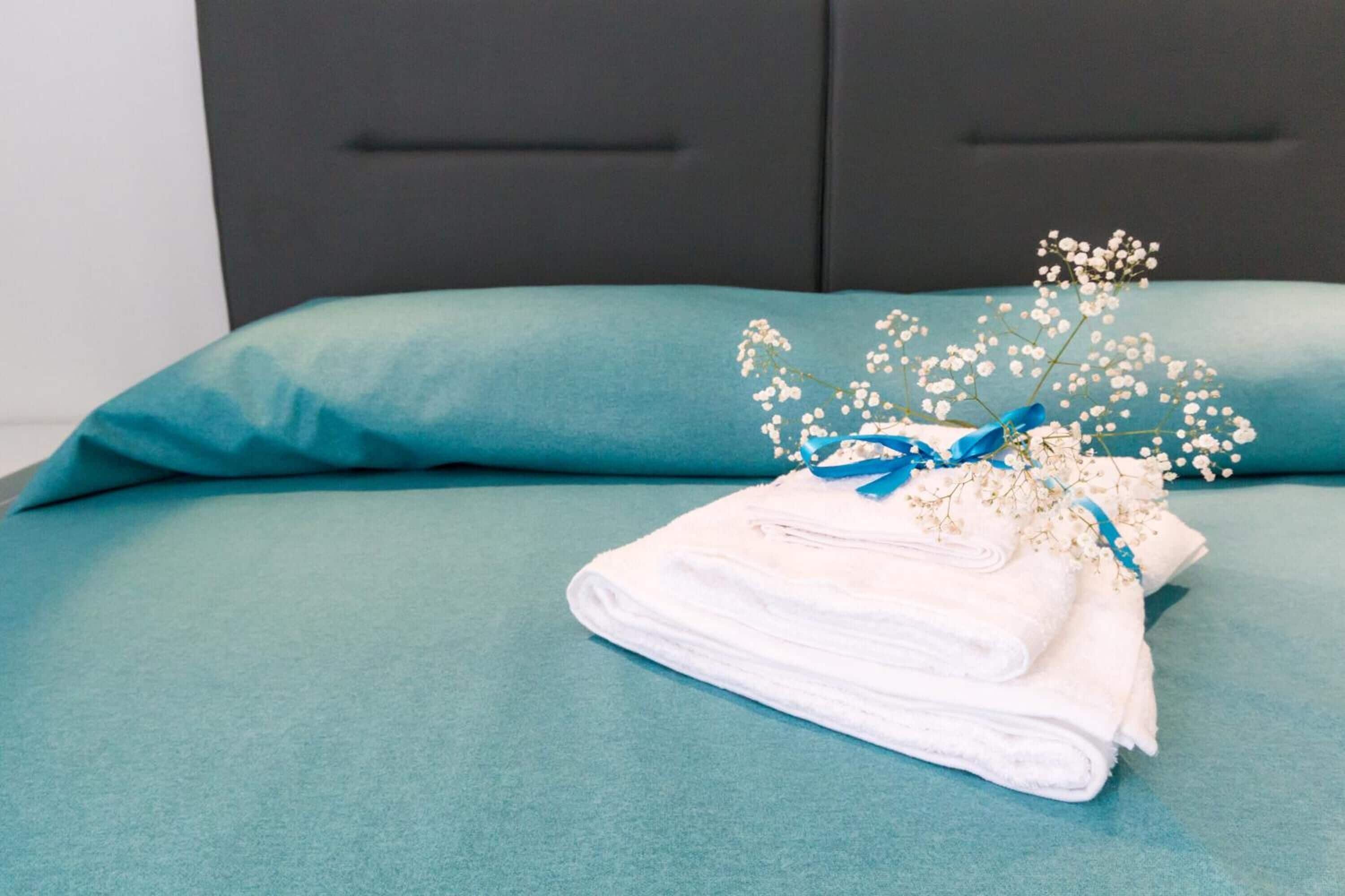 A neatly made bed with a teal bedspread and pillows, a stack of white towels, and a decorative arrangement of white flowers and blue ribbon.