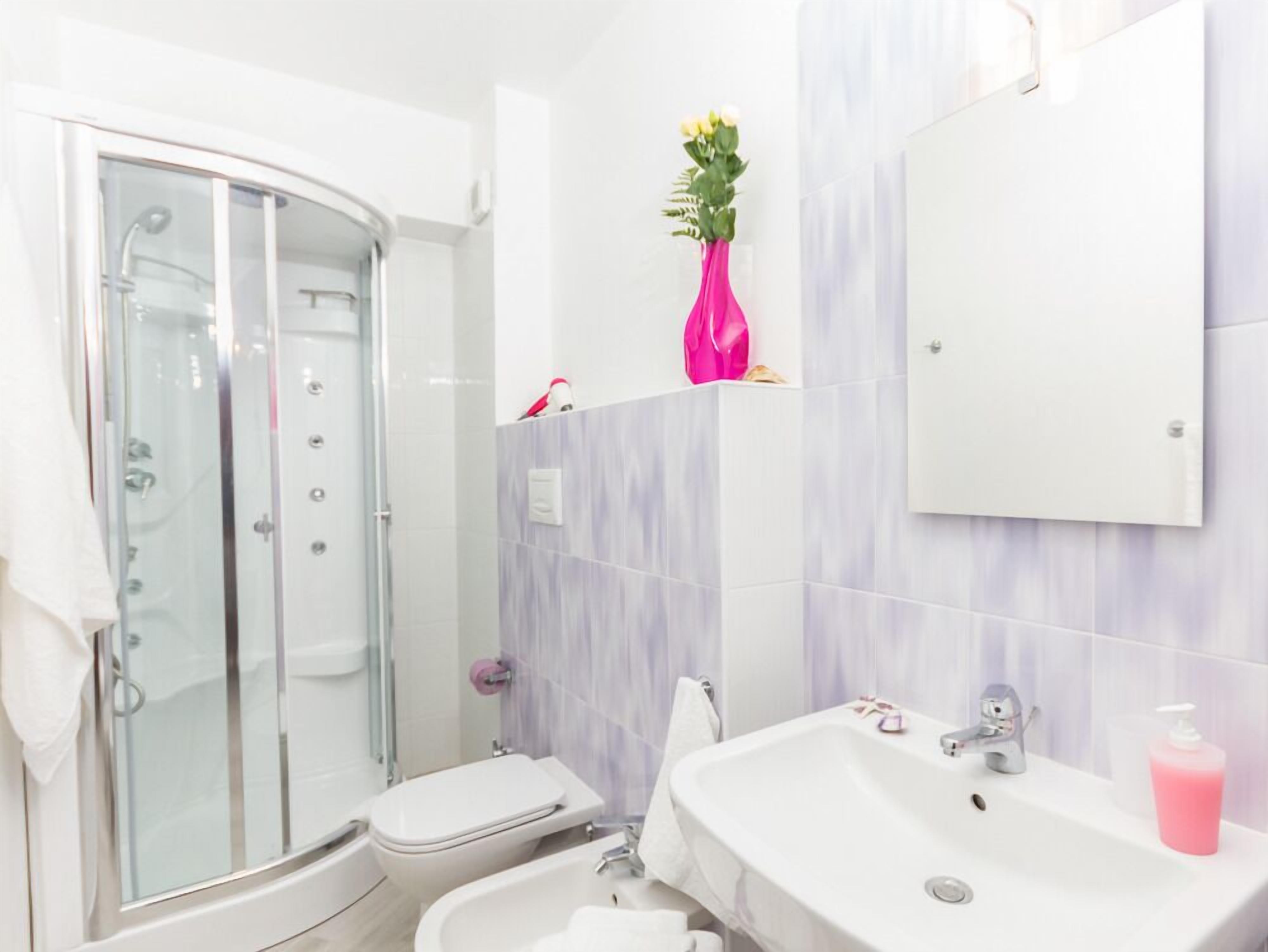 Une salle de bain moderne avec une douche à parois de verre, des toilettes blanches, un lavabo avec un miroir et un vase rose orné de fleurs, posé sur une étagère.