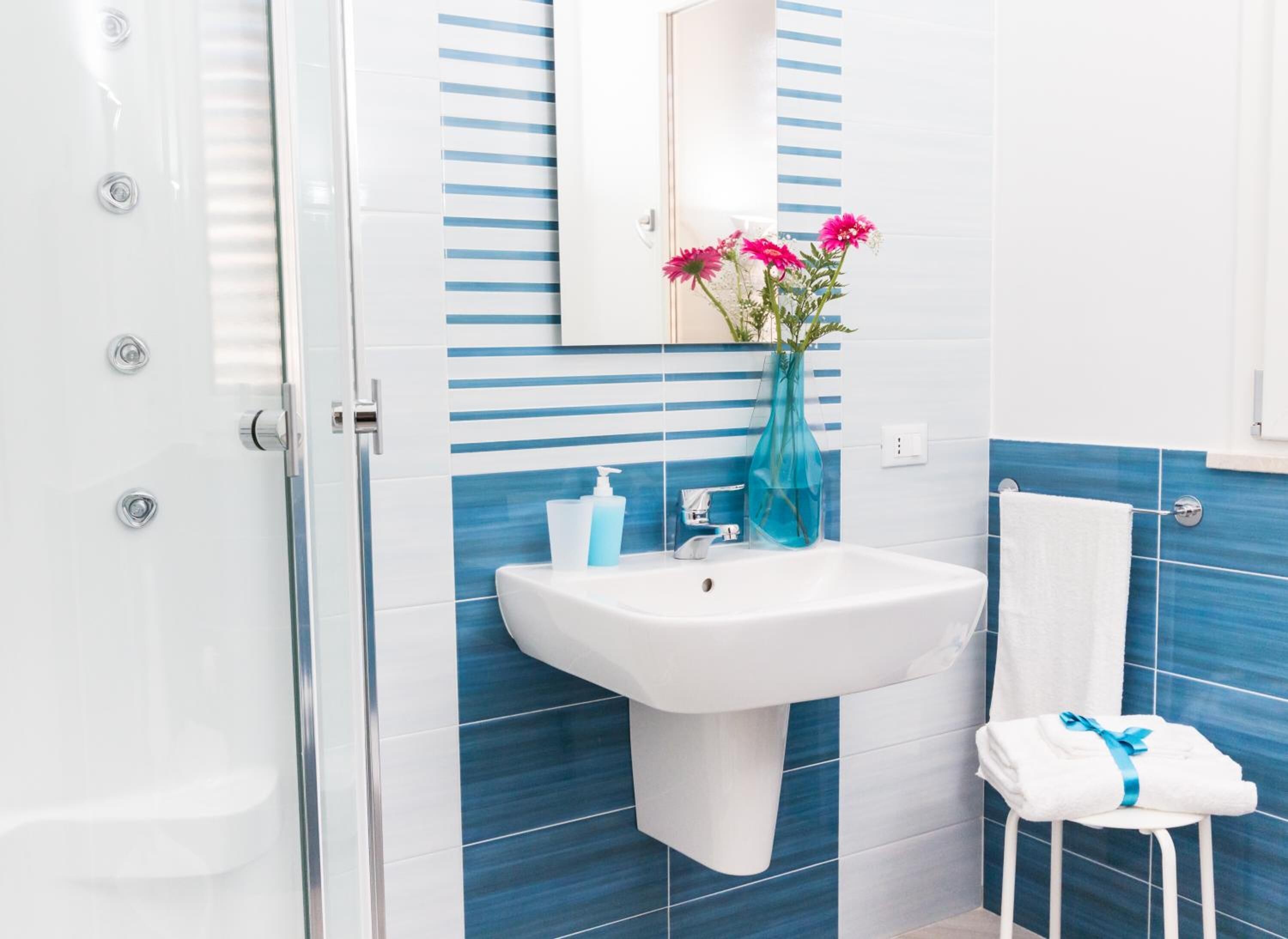 A bathroom with a white sink, blue-striped tiles, a glass shower enclosure, a white towel, and a blue vase with flowers.