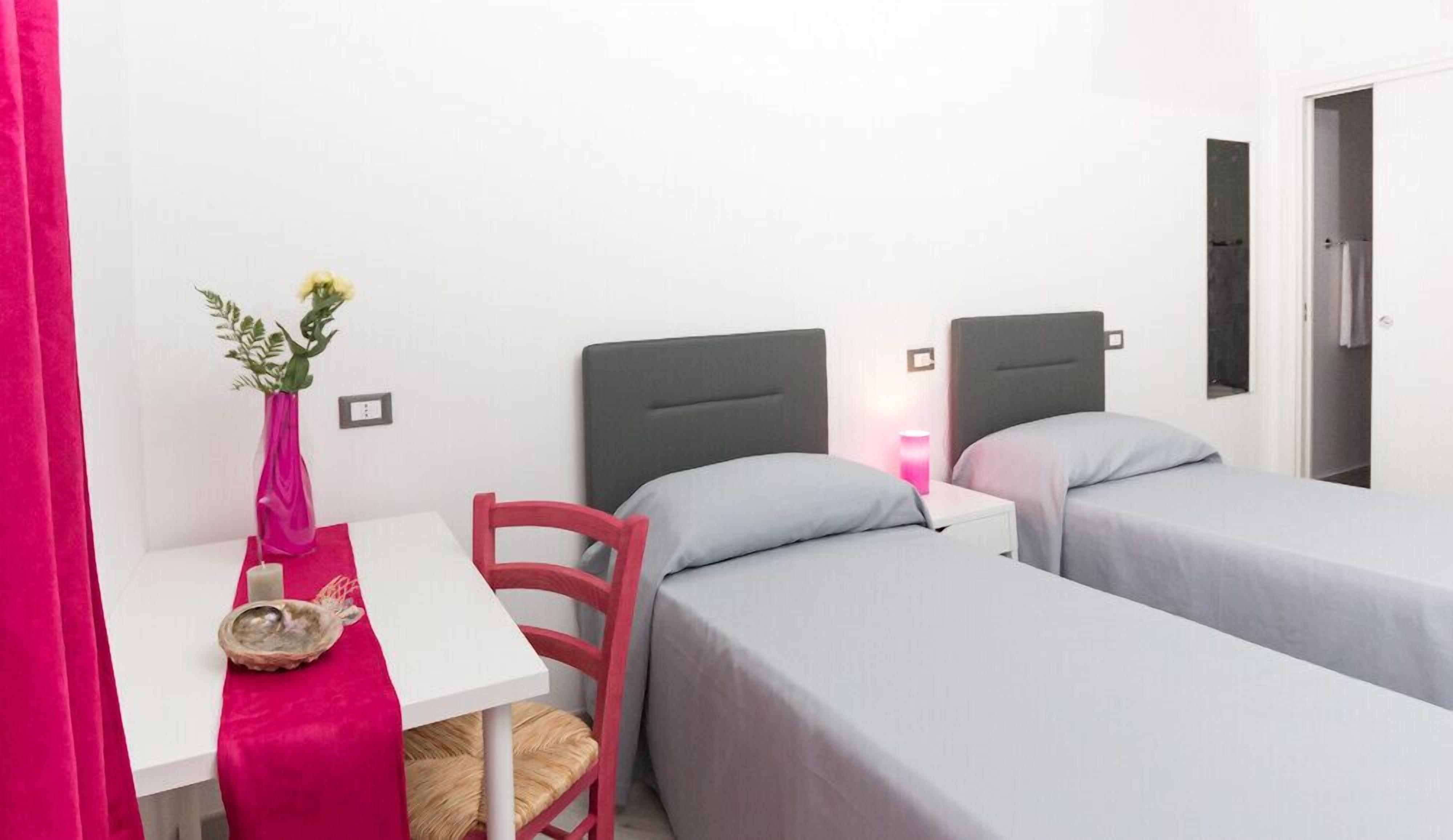 Ein Hotelzimmer mit zwei Einzelbetten, einem kleinen Schreibtisch mit Stuhl, einem rosa Vorhang und einer Blumenvase.