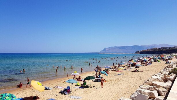 A crowded beach with people swimming and sunbathing, and a rocky shoreline.