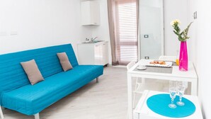 A modern, well-lit room with a blue sofa, a small white table with a plate of food and two glasses, and a pink vase with flowers.
