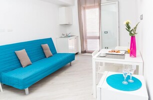 Una stanza moderna e luminosa con un divano blu, un piccolo tavolo bianco con un piatto di cibo e due bicchieri, e un vaso rosa con fiori.