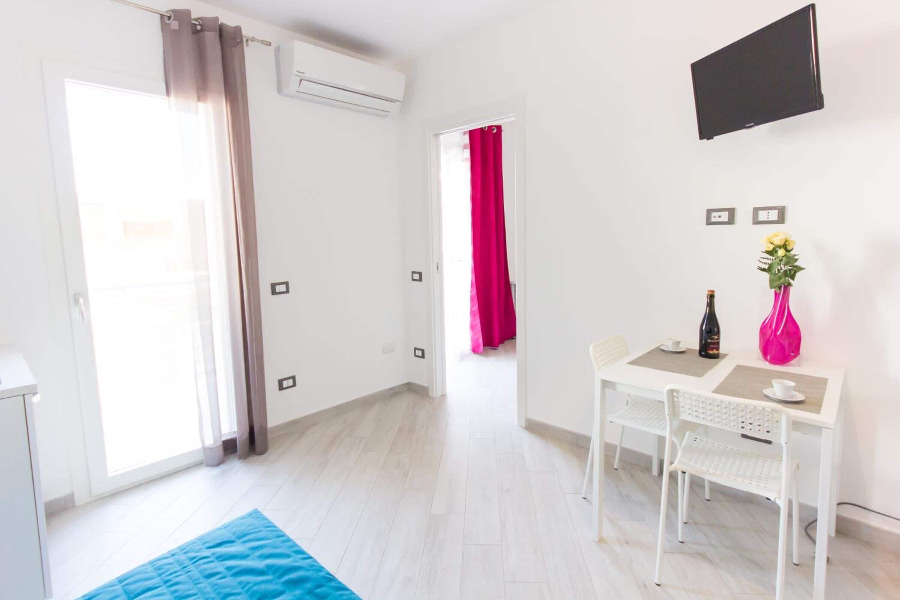 A bright, modern room with a white desk, two chairs, a flat-screen TV, and a window with curtains.