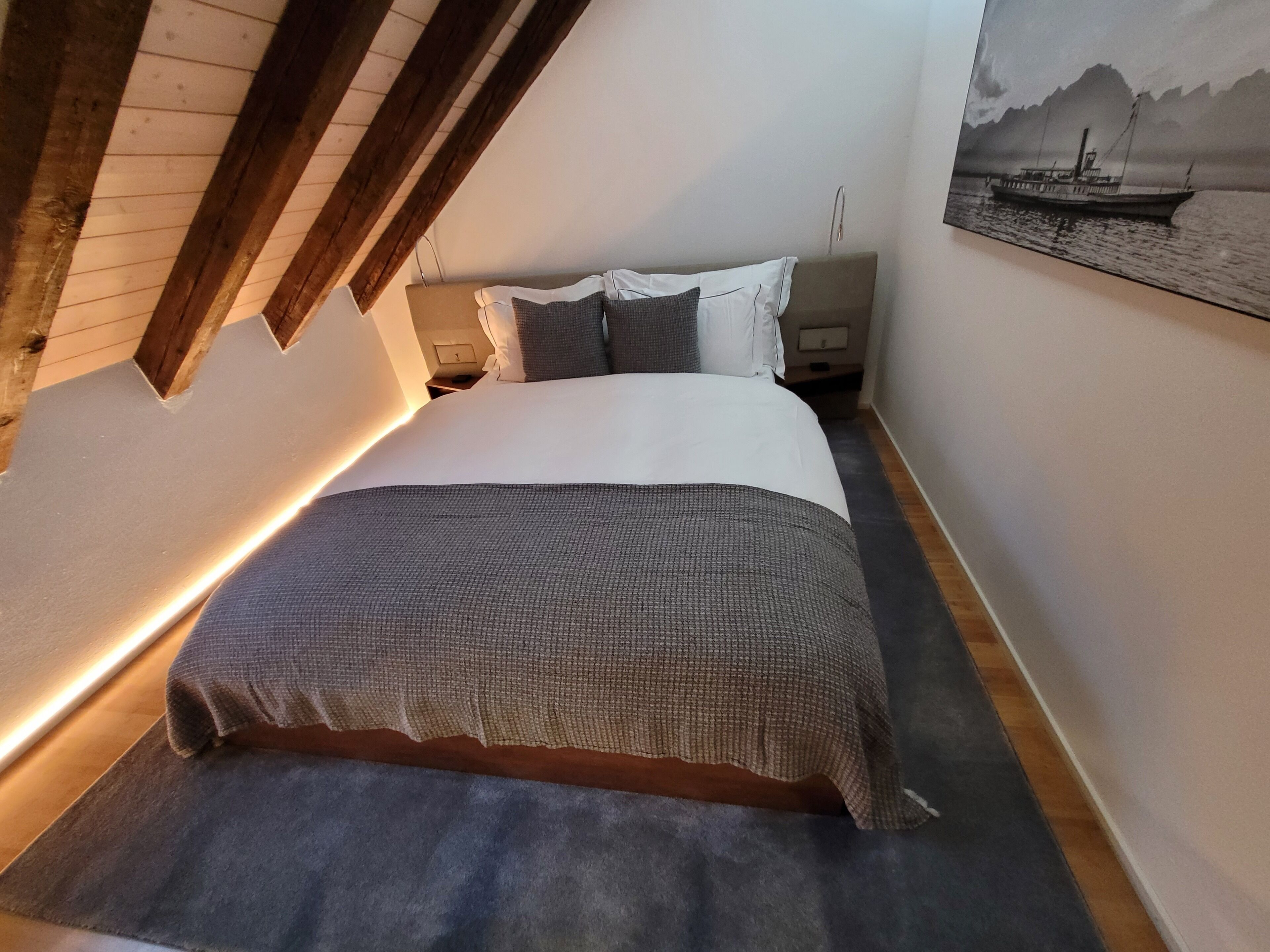 Une chambre avec un plafond en pente, un lit recouvert d’une couette grise et blanche, une table de chevet et une lampe fixée au mur.