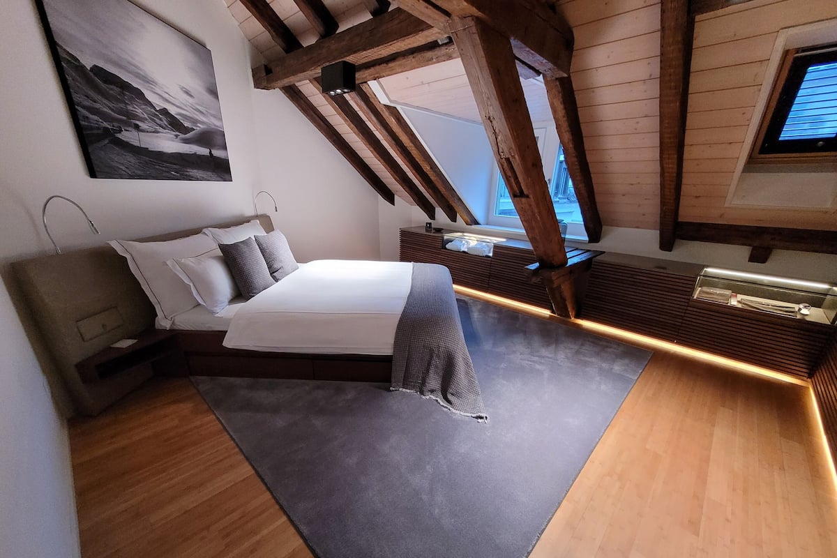 A bedroom with a bed, a nightstand, a wooden ceiling, and a window.