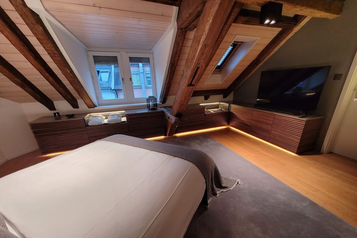 A bedroom with a bed, a TV, a window, and a wooden ceiling.