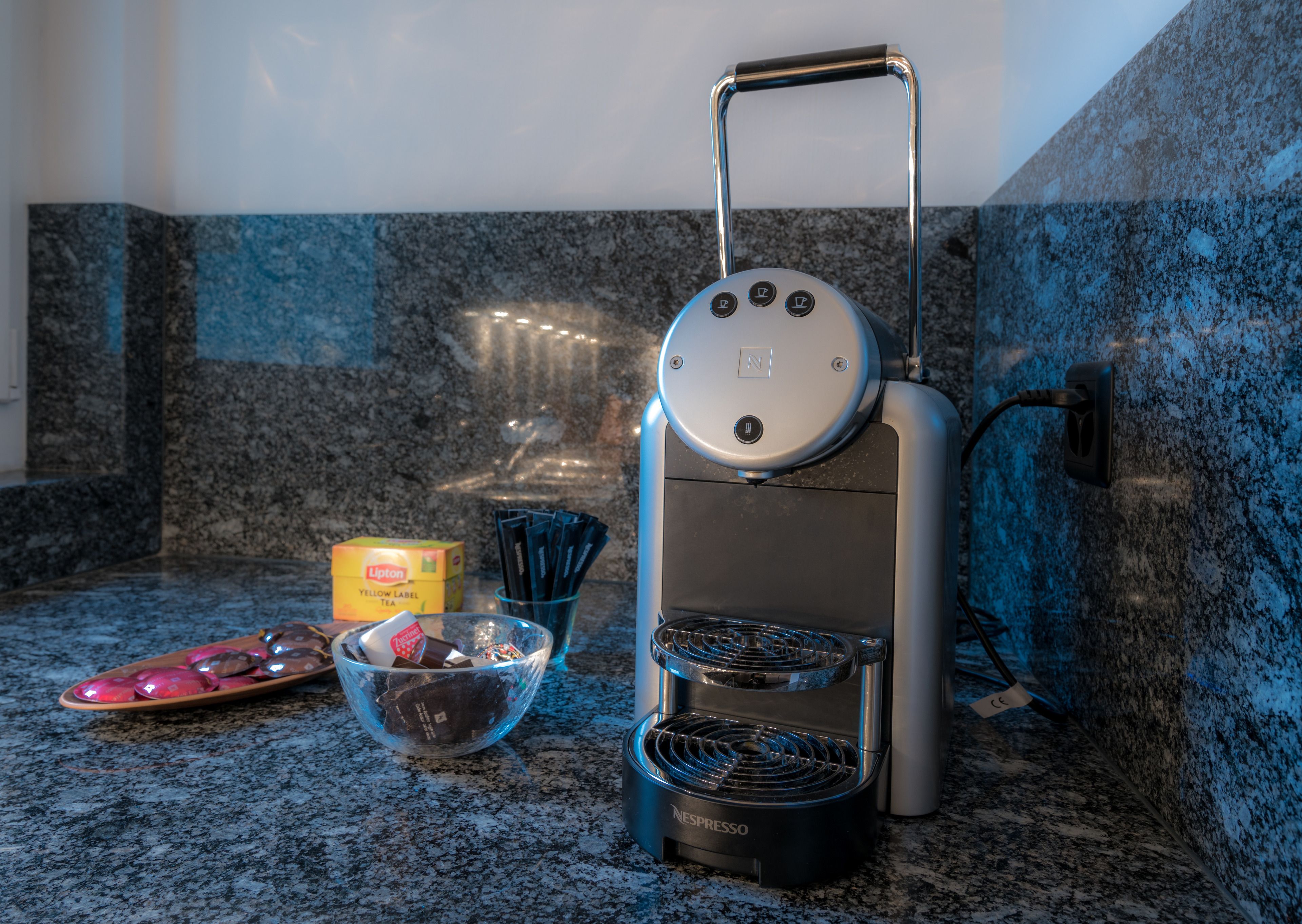 A Nespresso coffee machine on a granite countertop with a Lipton tea box and a bowl of chocolate.