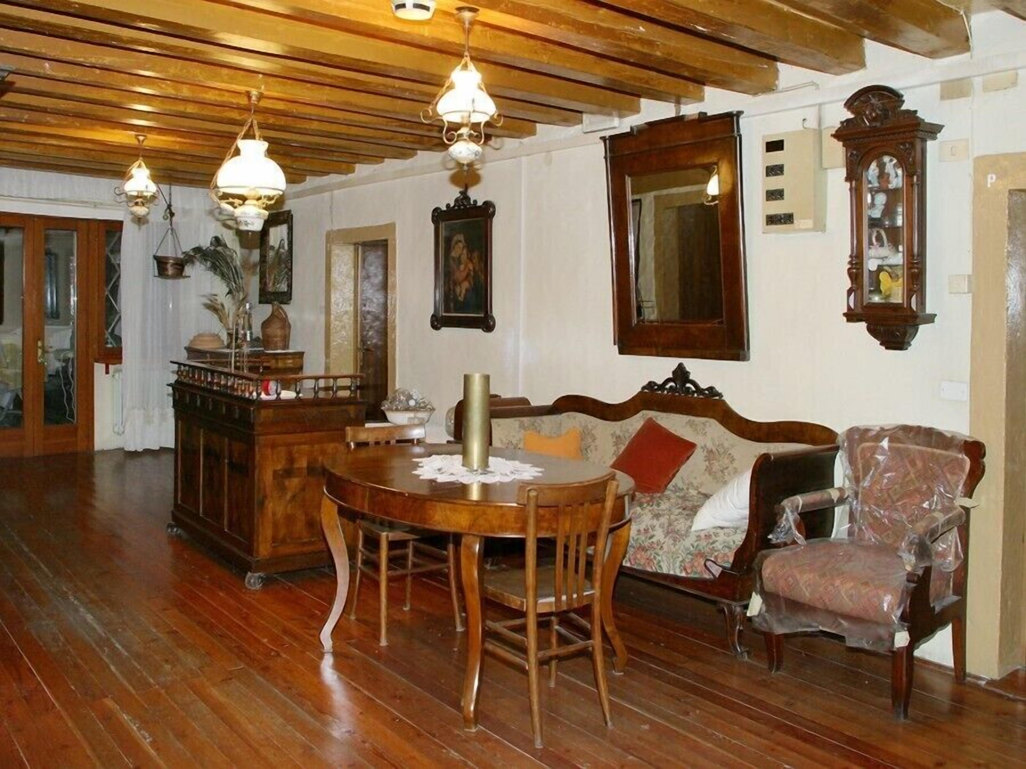 A wooden-floored room with a dining table, chairs, a sofa, and a chair with a plastic cover. There are wall-mounted lights and a mirror.