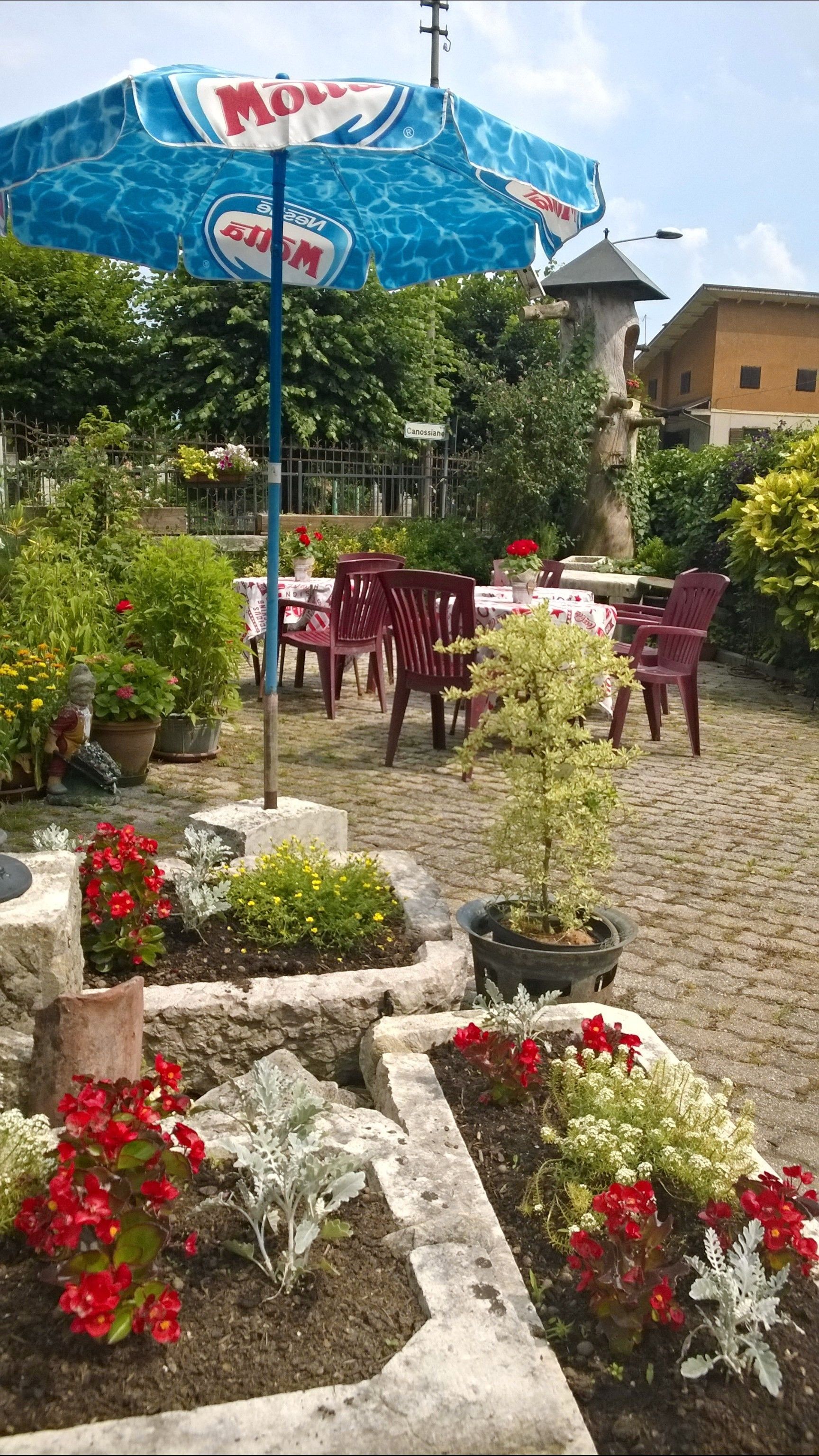 An outdoor patio area with a blue umbrella, stone planters with flowers, and red plastic chairs.