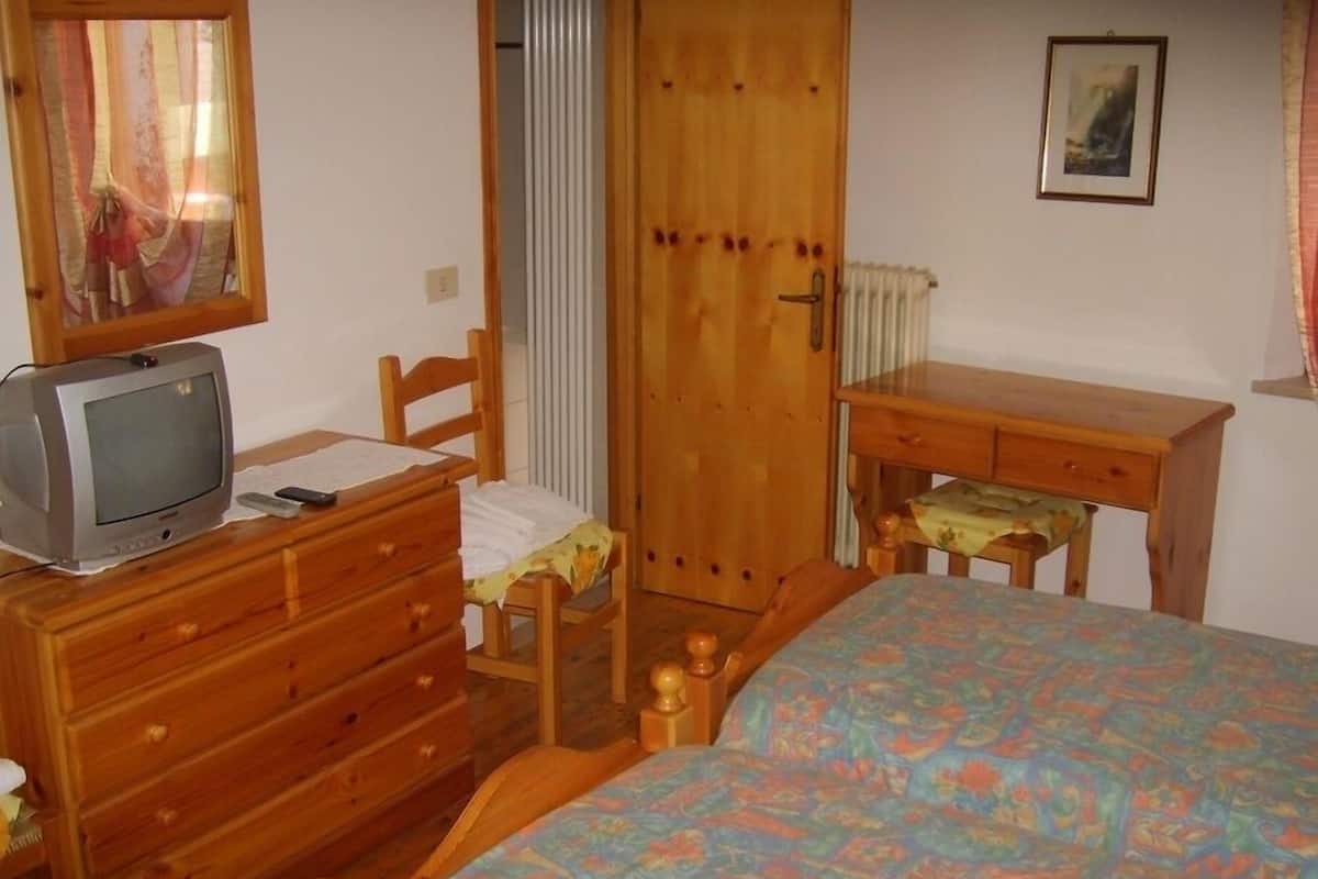 Een slaapkamer met een houten dressoir, een bed met een kleurrijk sprei, een klein houten bureau, een televisie en een deur met een raam.