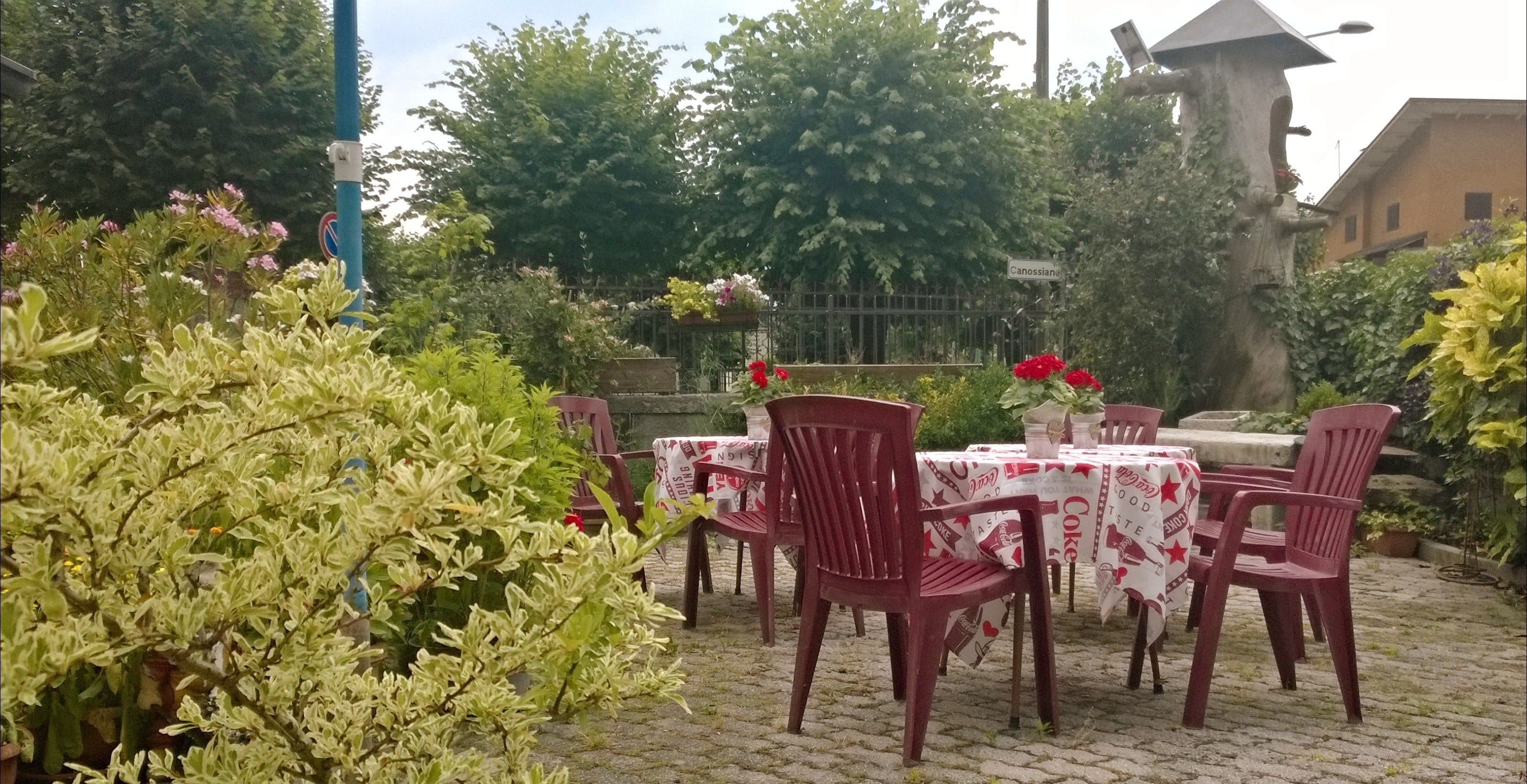 An outdoor seating area with tables and chairs, surrounded by greenery and a metal fence.
