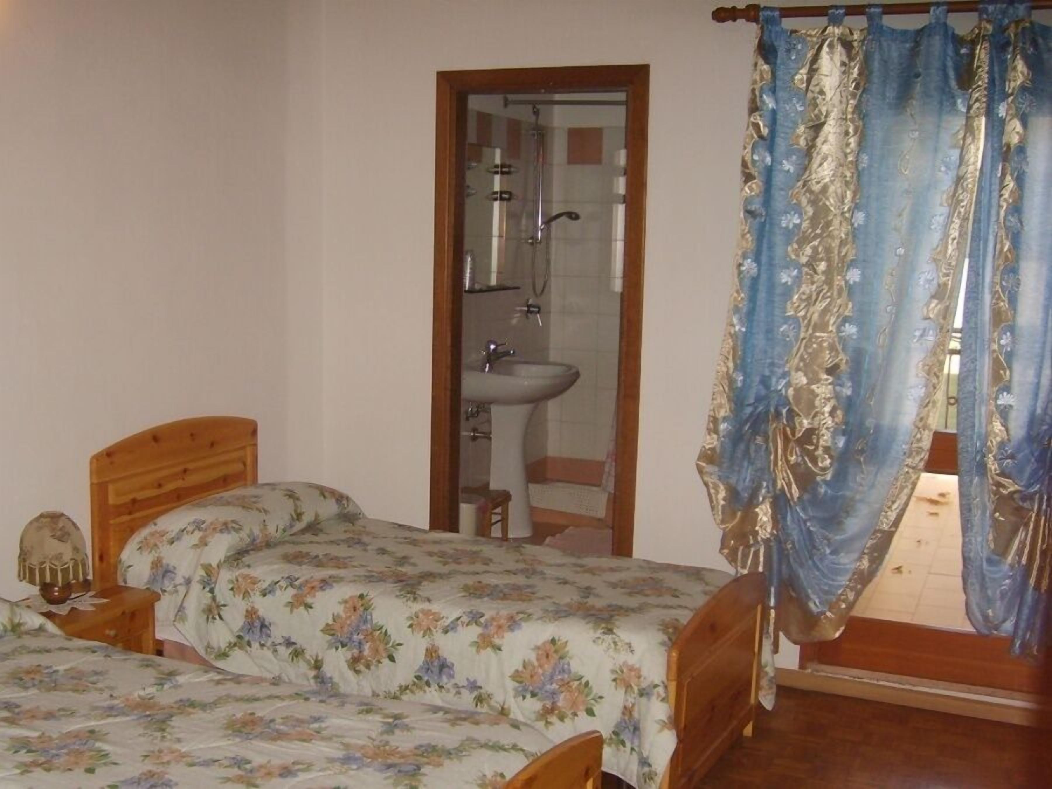 Ein Schlafzimmer mit zwei hölzernen Betten, einem Nachttisch aus Holz, einem Spiegel, einem durch eine Glastür einsehbaren Badezimmer und einem Fenster mit blauen Vorhängen.