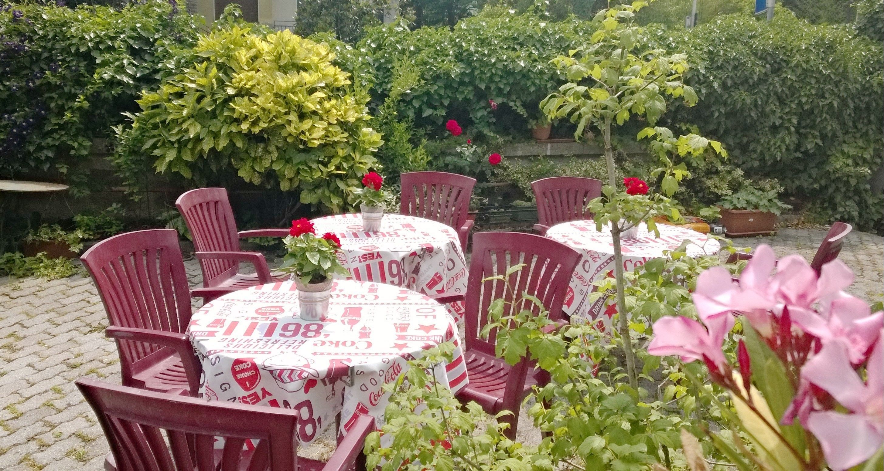 Zona salotto all'aperto con tavoli rotondi ricoperti da una tovaglia a tema Coca-Cola, circondata da sedie in plastica rossa.
