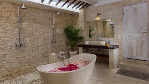 A modern bathroom with a freestanding bathtub filled with rose petals, a stone wall, a large mirror, and a shower area. - Villa Dewi (Seminyak)