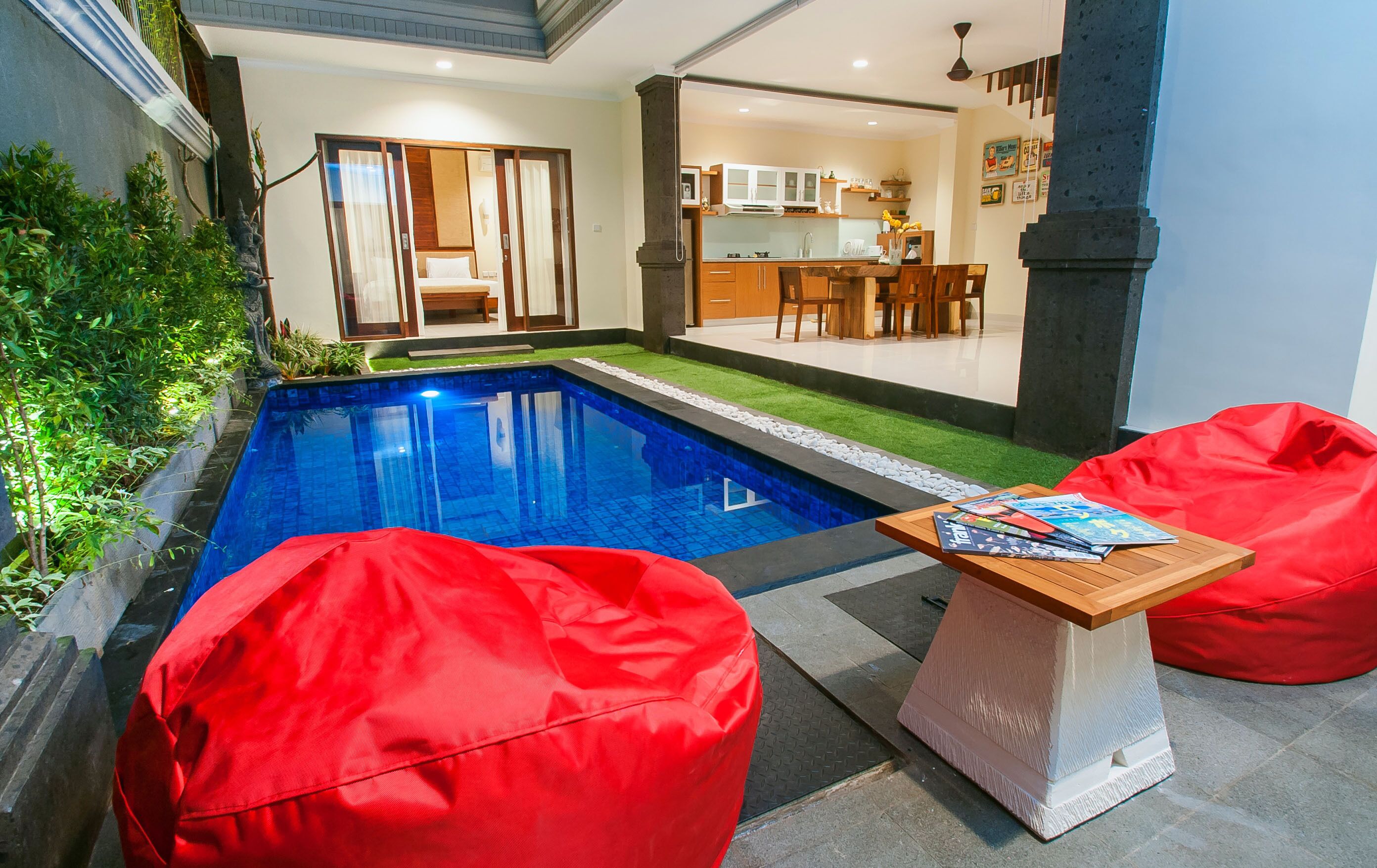 Una moderna area piscina con pouf rossi e un tavolo in legno, una camera da letto con porta scorrevole e una cucina visibile attraverso una porta aperta.