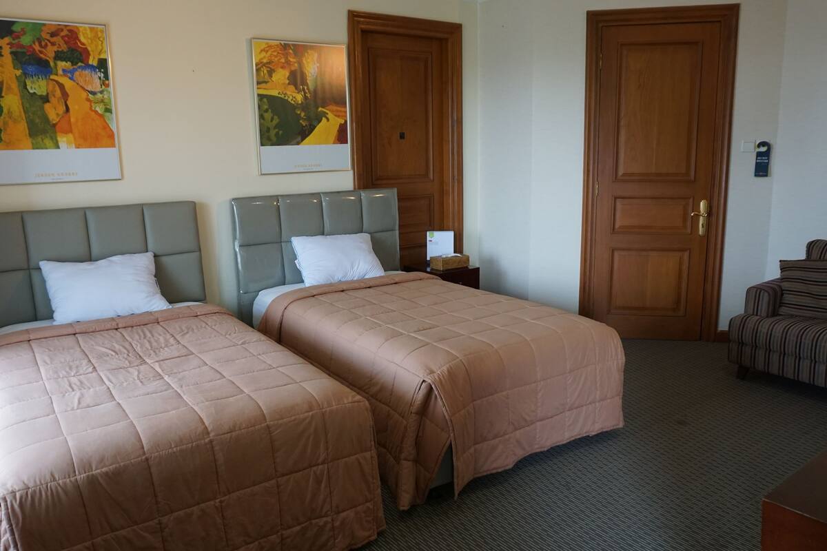 Una habitación de hotel con dos camas, una puerta de madera, un sillón a rayas y dos cuadros en la pared.