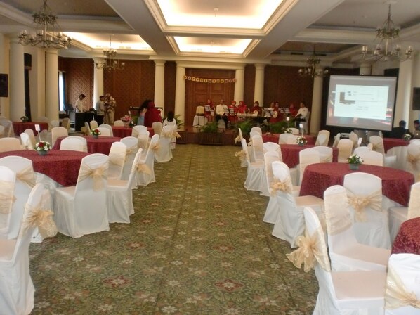 Un salón de banquetes con filas de mesas cubiertas con manteles blancos y sillas con fundas blancas y cintas doradas.