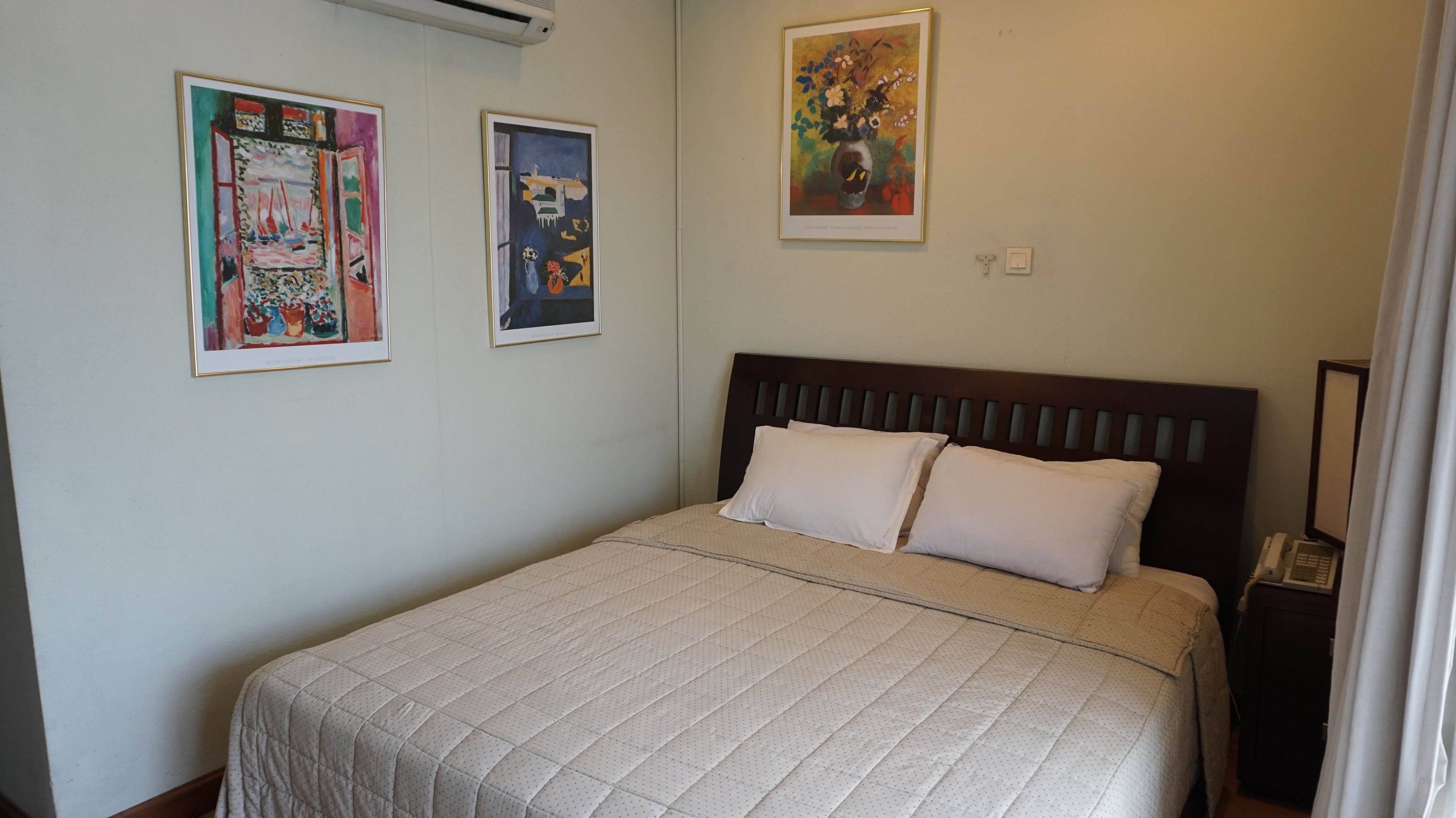 Una cama bien hecha con cubrecama acolchado, cabecera de madera y dos almohadas. Se observan dos obras de arte enmarcadas en la pared sobre la cama.