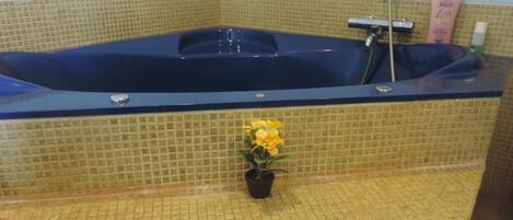 Una bañera con interior azul, una flor amarilla en maceta y un muro cubierto de baldosas amarillas.