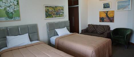 A hotel room with two beds, a wardrobe, a green armchair, and several paintings on the walls.