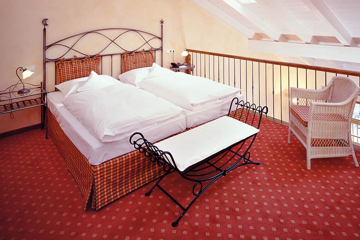 A double bed with a metal frame and checkered bedding, a small table, a chair, and a nightstand with a lamp.