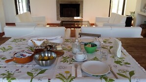 Una mesa de comedor con mantel floreado, diversos alimentos y cubiertos, en una habitación con sofás blancos y chimenea.