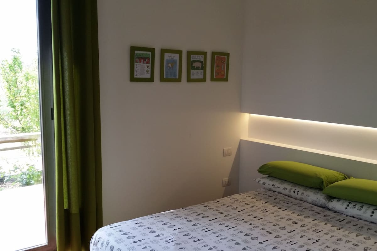 Ein Schlafzimmer mit Bett, grünen Kissen, einem Nachttisch und einem Fenster mit Vorhängen. An der Wand hängen vier gerahmte, lehrreiche Poster.
