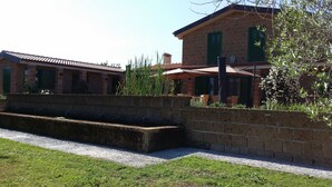 Un edificio de ladrillo con contraventanas verdes, un espacio de estar al aire libre cubierto y una pequeña fuente de agua.