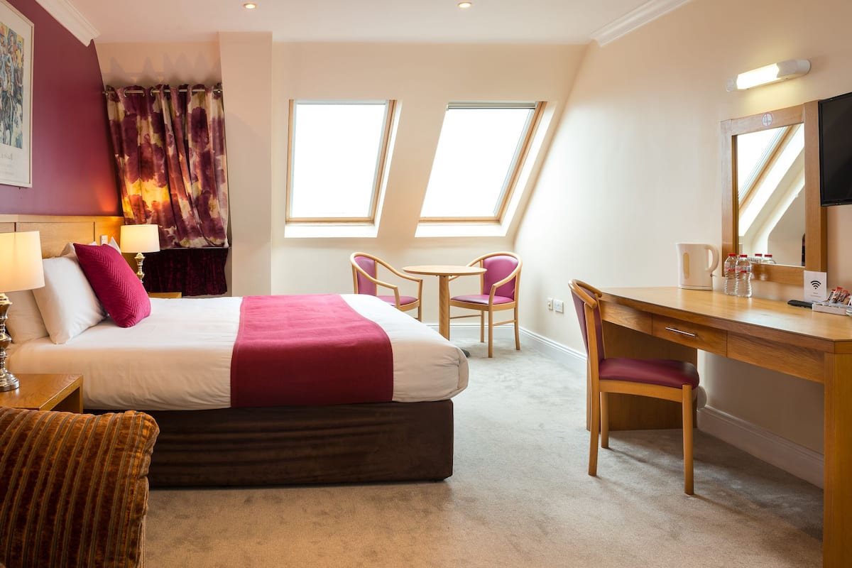 A hotel room with a bed, desk, two chairs, a small table, a mirror, and two windows.
