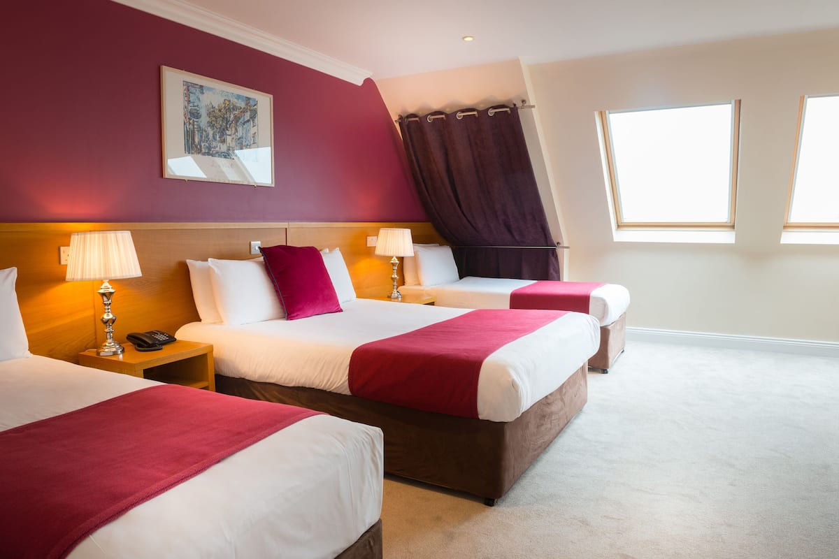 A hotel room with two beds, a painting on the wall, a window with curtains, and a lamp on the nightstand.