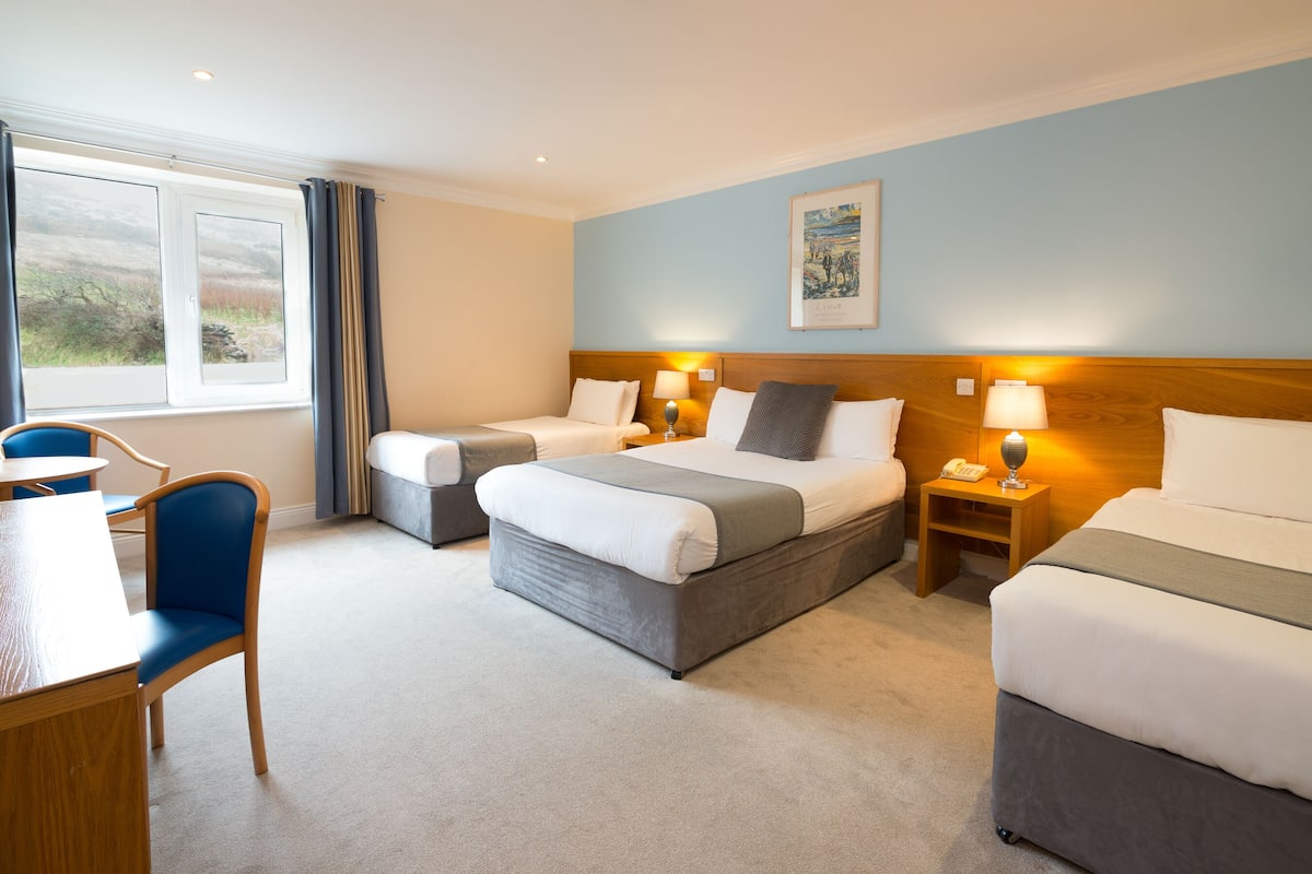 A hotel room with two beds, a desk, and a chair. There is a window with curtains, a painting on the wall, and a lamp on the bedside table.