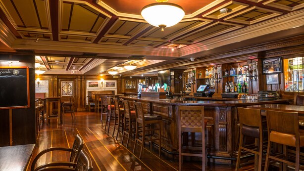 Zona de bar con suelo de madera, taburetes y una barra con varias botellas y una caja registradora.