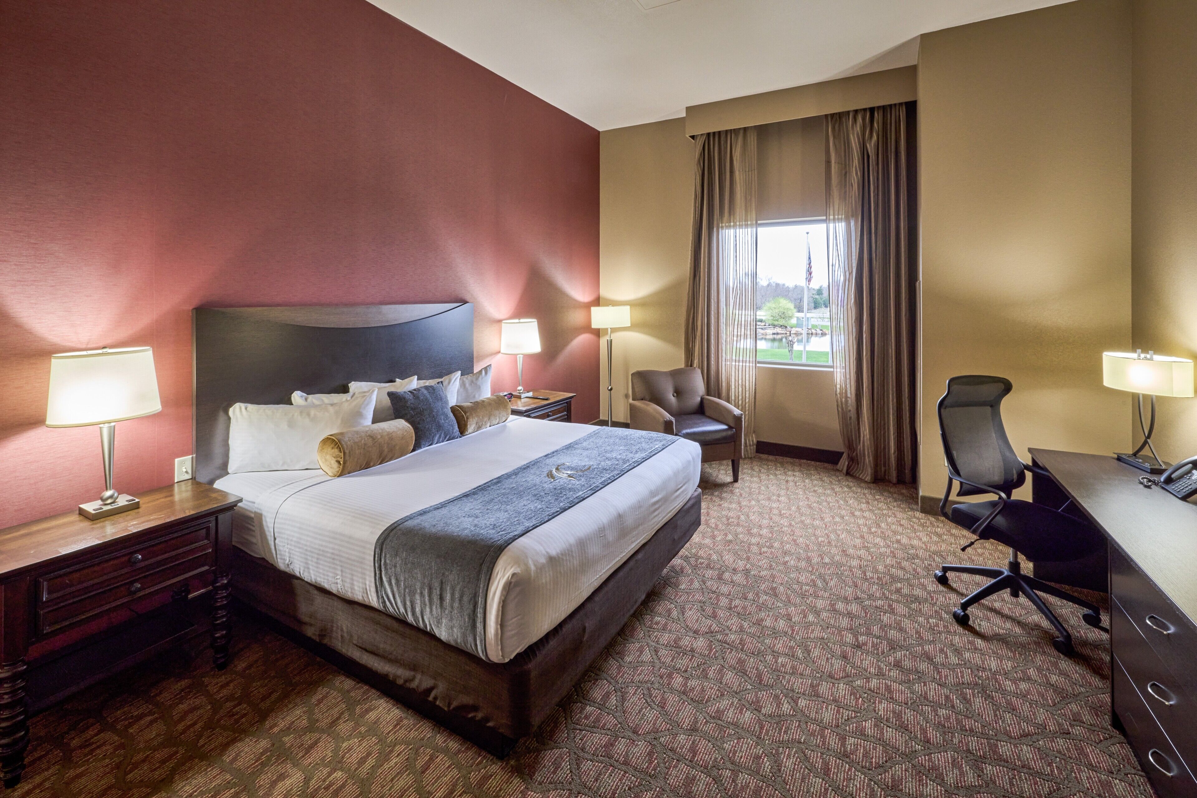 A hotel room with a large bed, two bedside tables with lamps, a desk with a chair, and a view of a park through the window.