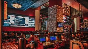 A casino with multiple gaming tables and chairs, a large screen displaying a game, and a stone wall with a logo. - Choctaw Casino & Resort Pocola, Trademark Coll by Wyndham (Pocola)