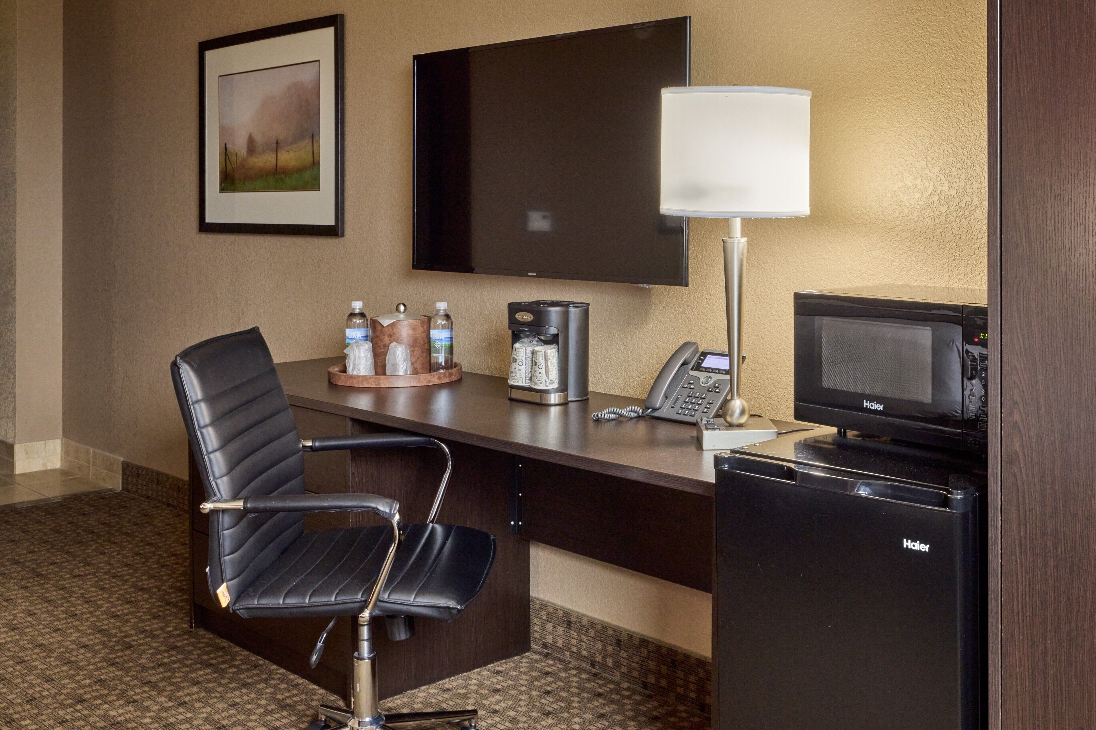 A hotel room with a desk, a chair, a flat-screen TV, a coffee maker, a microwave, and a refrigerator.
