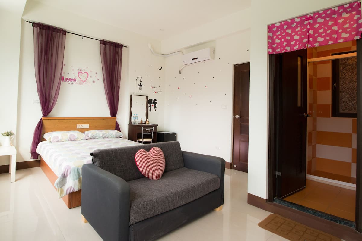 A bedroom with a bed, a bedside table, a grey sofa with a heart-shaped pillow, a desk with a chair, and a wardrobe with a pink heart-patterned curtain.