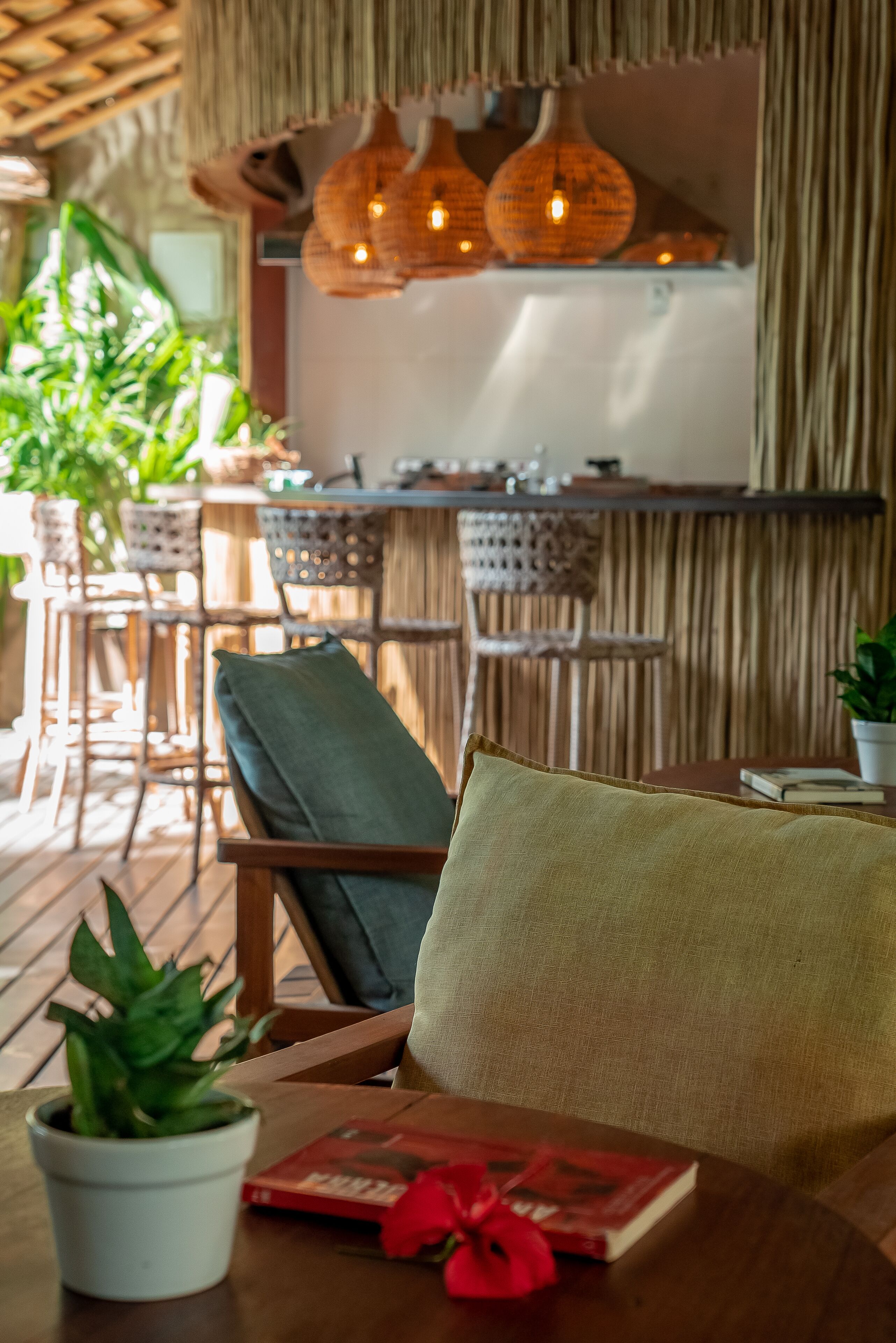 Uma área de estar acolhedora, com mobiliário de madeira, uma mesa com um livro e uma flor vermelha, e luminárias pendentes.