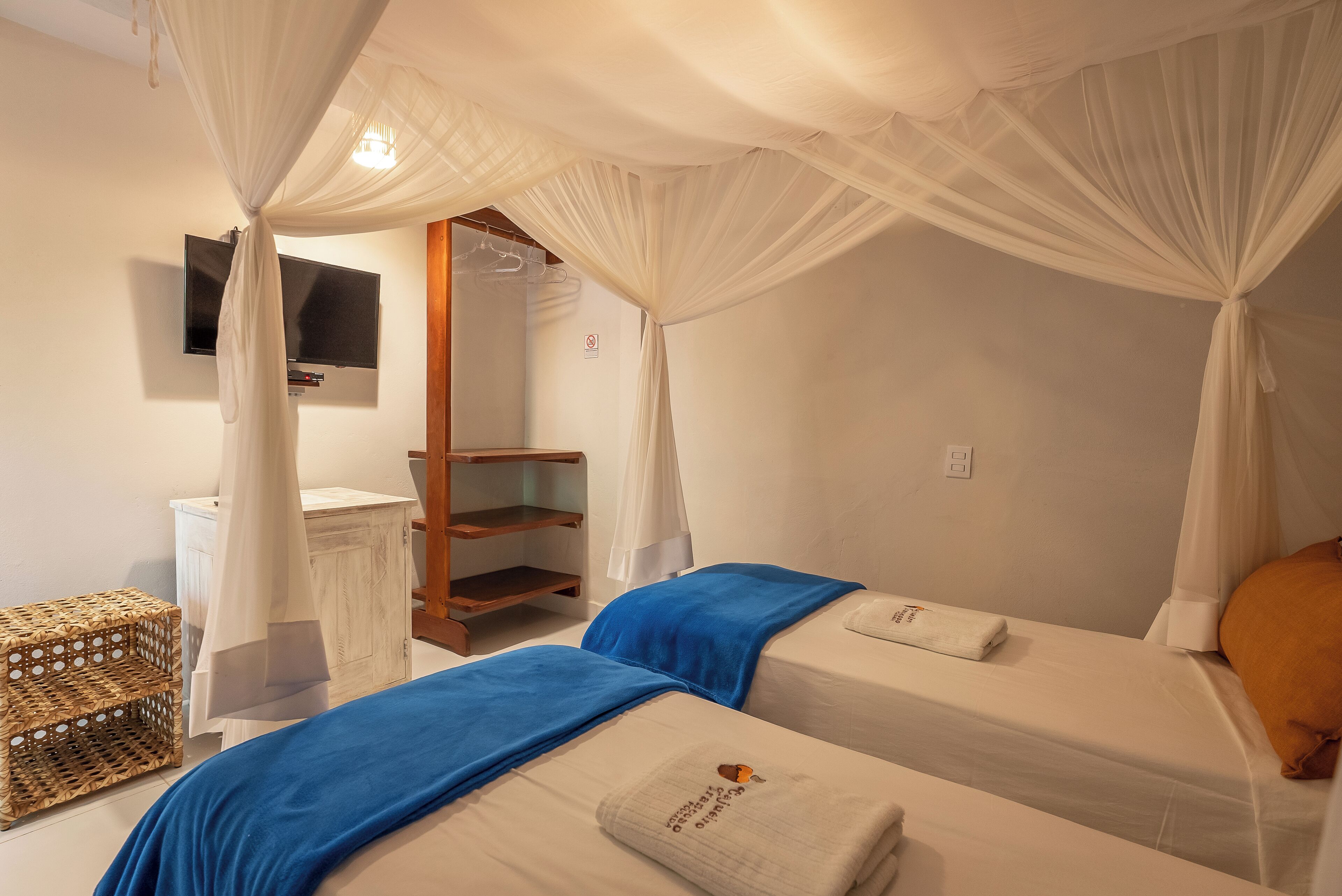 Habitación de hotel con dos camas, un televisor instalado en la pared, un mueble de madera y un baúl de mimbre.